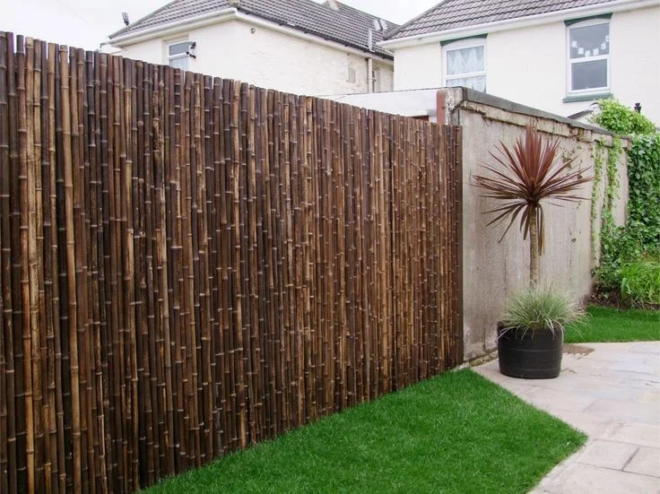 Vertical rolls of natural black bamboo fencing poles screening a backyard garden border next to a small green grass lawn.