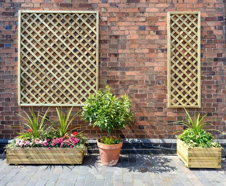 Two light wood diagonal lattice trellis panels mounted on a rustic red brick wall above wooden planter boxes and a potted topiary tree.