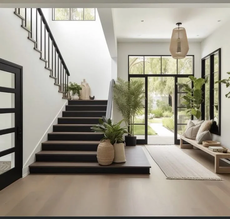 A bright, spacious modern Apartment entryway featuring a dark wood staircase with a black metal railing on the left. Large black-rimmed glass doors and windows open up to a lush green backyard, filling the space with natural light. A wooden bench with throw pillows sits on the right under a large window, complemented by a light-colored runner rug on the light oak floors. Potted plants, including a fiddle-leaf fig and ferns, add vibrant greenery alongside woven basket planters and a woven pendant light hanging from the high ceiling.