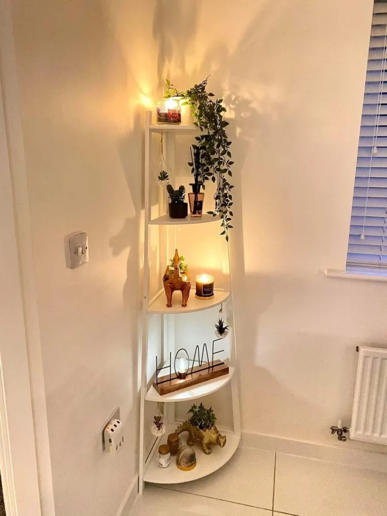 A five-tier white corner ladder shelf stand decorated with glowing scented candles, a trailing artificial ivy plant, small succulents, a wire 'HOME' sign, and gold animal figurines in a warm, warmly lit room.