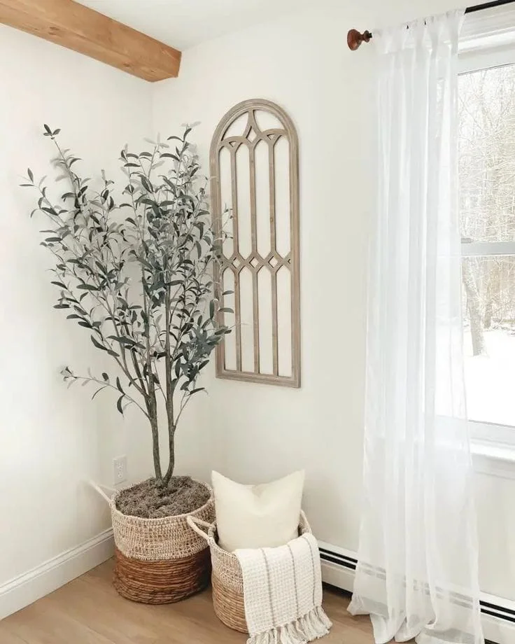 Cozy Farmhouse Corner Decor Ideas with Olive Trees A bright room corner styled with a potted faux olive tree in a woven basket, a decorative arched wooden window frame on the wall, and a smaller basket holding a white throw pillow and blanket next to a window with sheer white curtains.