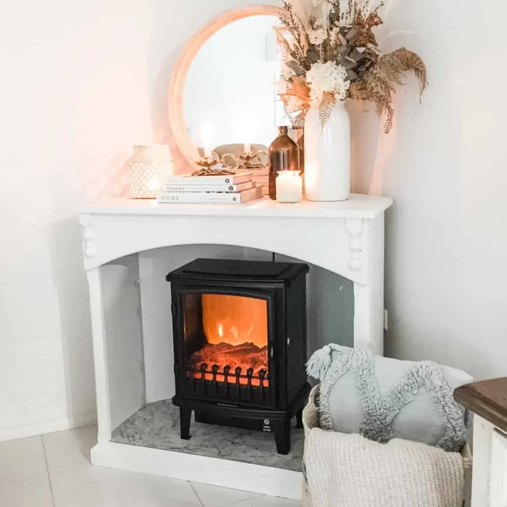 How to Create a Cozy Faux Fireplace Corner in Any Room A cozy room corner featuring a freestanding black electric fireplace inside a white faux mantel, topped with a round mirror, glowing candles, and a vase of dried flowers.