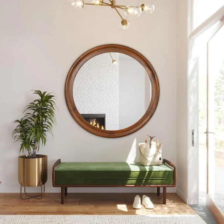 A mid-century modern entryway features a green velvet bench with a dark wood frame underneath a large round wooden mirror. A canvas tote bag sits on the bench with white sneakers tucked underneath, while a tall gold planter stands to the left. The bright space is illuminated by a gold sputnik chandelier against clean white walls and light hardwood floors.