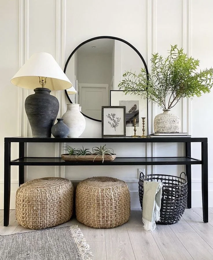 A modern entryway features a long black two-tiered console table underneath a large round black-framed mirror against a white paneled wall. The tabletop is styled with a large black ceramic lamp, textured stoneware vases, framed artwork, and green branches, while a small dough bowl with air plants sits on the middle shelf. Two woven jute poufs and a black wire basket with a throw blanket rest neatly on the floor underneath.