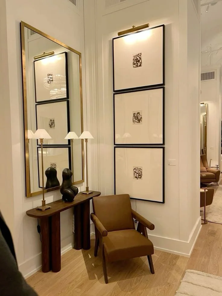 A high-end entryway corner features three square black-framed abstract art pieces stacked vertically beneath a gold picture light. To the left, a tall brass-framed mirror hangs above a narrow dark wood console table styled with two slender buffet lamps and a black contemporary sculpture. A mid-century brown leather armchair sits comfortably in the corner over light hardwood flooring.