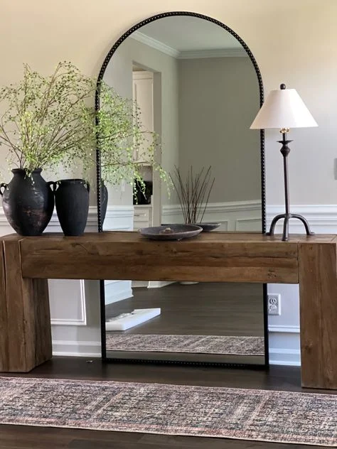 A rustic entryway features a thick, chunky wooden console table set in front of a massive, full-length arched floor mirror with a beaded black border. The table is styled with two large, textured black ceramic vases holding delicate green branches, a central decorative bowl, and a thin dark metal table lamp. A vintage-style patterned runner rug rests on the dark wood floor in front.