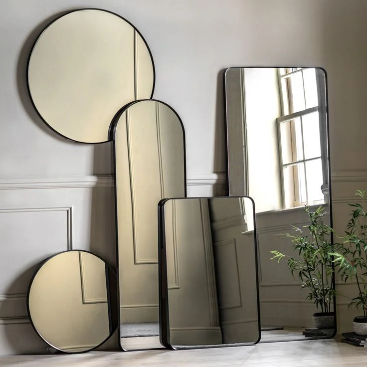 A collection of modern mirrors with sleek black metal frames is clustered against a neutral gray wall with traditional molding. The arrangement features various geometric shapes, including two round mirrors, a tall arched floor mirror, and two rectangular mirrors with rounded corners. Leaning against the wall at varying heights, the mirrors reflect the indoor space and a small potted green plant.
