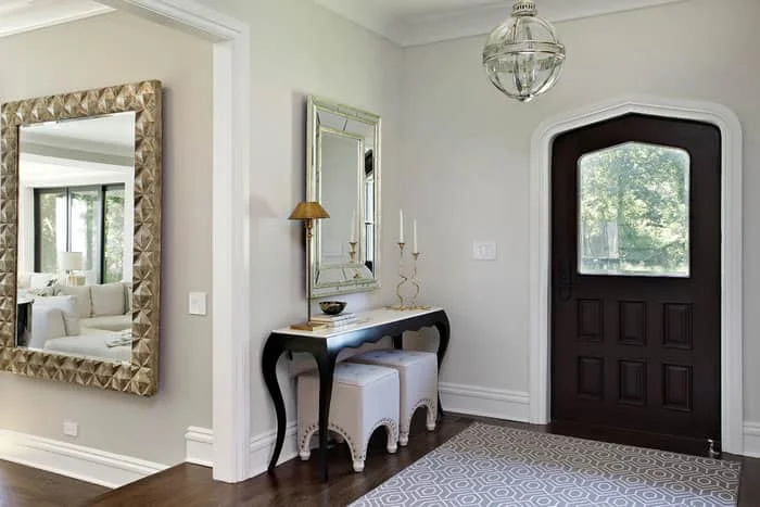 An elegant, bright home foyer features a dark wood front door with an arched frame and an overhead glass globe light. A narrow black console table with a marble top sits against a light gray wall, styled with a small gold lamp and candlesticks beneath a silver mirror. Two white upholstered ottomans are tucked underneath, while a massive textured gold-framed mirror on the adjacent wall reflects a living room.