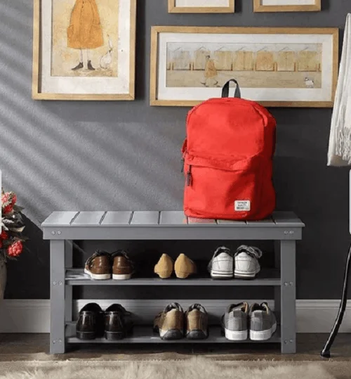A 2-tier gray wooden shoe rack bench in an entryway with shoes organized on the shelves, a bright red backpack sitting on top, and framed artwork hanging on a dark gray wall.