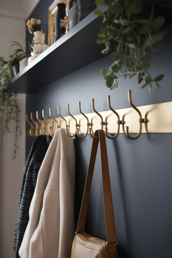 Modern Brass Wall Hooks on Navy Accent Wall A close-up of a long, metallic gold or brass coat rack with multiple curved hooks mounted on a dark navy blue entryway wall. A cream coat, plaid jacket, and brown leather purse hang from the hooks below a black floating shelf adorned with green plants.