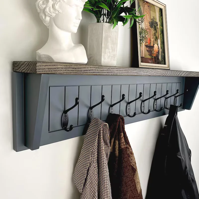 Grey Wall-Mounted Coat Rack with Wood Top Shelf A modern grey wall-mounted coat rack featuring a natural wood shelf and a row of black metal hooks. A houndstooth blazer and a black leather jacket hang from the hooks, while the top shelf displays a white classical bust sculpture, a potted green plant, and a framed art piece.