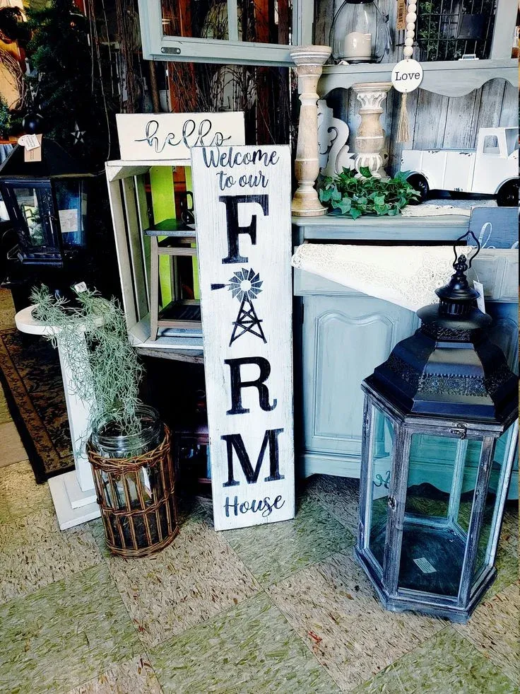 A rustic, white-painted wooden vertical sign reading "Welcome to our FARM House" with a windmill design, displayed alongside vintage lanterns, green foliage, and blue-grey wooden furniture.