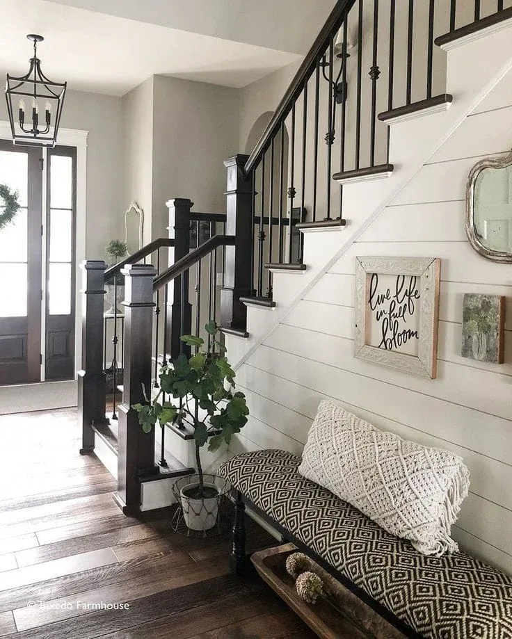 A modern farmhouse foyer featuring a dark wood staircase with black metal spindles, a white shiplap accent wall, an upholstered geometric print bench with a textured lumbar pillow, and a potted plant.