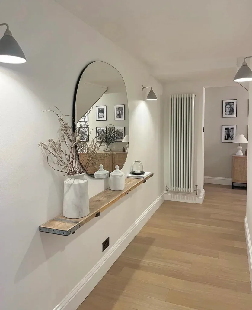 Modern Minimalist Hallway with Large Oval Mirror and Floating Shelf A bright minimalist hallway featuring a large oval mirror mounted above a rustic floating wooden shelf. The shelf holds a white marble vase with dried branches, while modern wall sconces illuminate the white walls and light oak flooring.