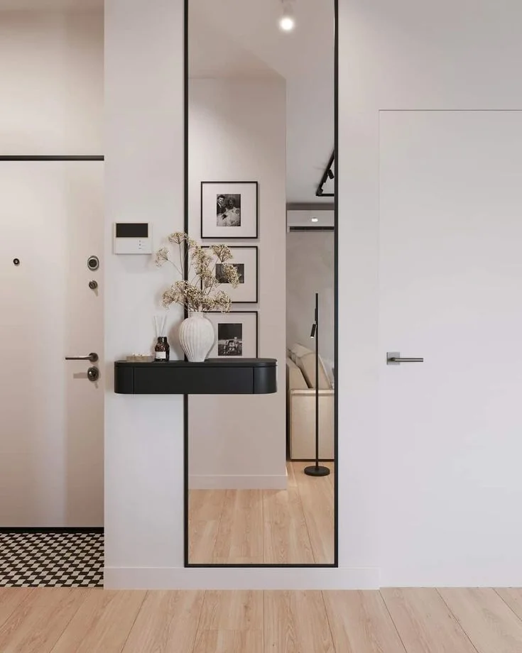 small hallway with Full Length Mirror and Black Floating Shelf A small entryway featuring a tall, thin full-length mirror with a slim black frame. A sleek black floating console shelf is mounted across the mirror, styled with a ribbed white vase of dried flowers and a reed diffuser against a clean white wall.