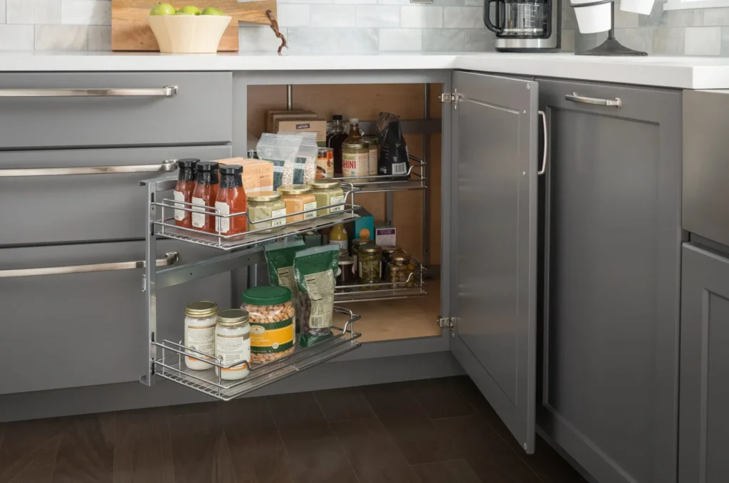 blind corner cabinet pull out wire organizer A grey lower kitchen corner cabinet open to reveal a two-tier metal wire pull-out shelf organizer filled with assorted pantry jars, sauces, and snacks.
