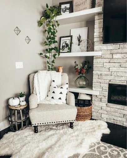 cozy living room corner decor with armchair and built in shelves Cozy living room corner featuring a beige upholstered armchair with a black and white geometric pillow, sitting on a white faux fur rug next to a stone fireplace and built-in floating shelves styled with plants and framed art.