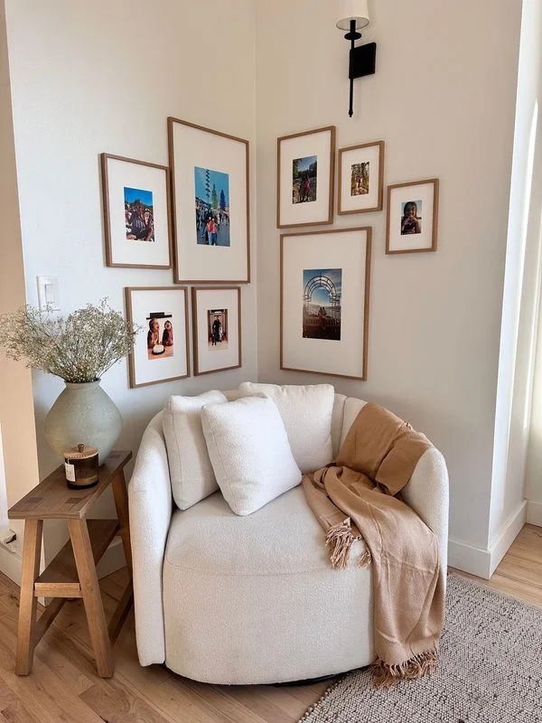 cozy living room corner with white boucle chair and corner gallery wall A cozy living room corner featuring a round white bouclé swivel armchair styled with a tan fringed throw blanket, nestled under a symmetrical corner gallery wall of light wood-framed photos.