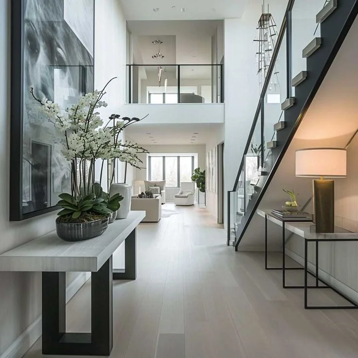A luxurious contemporary foyer featuring a double-height ceiling, light wood flooring, and a floating staircase with glass railings. A minimalist white console table displays white orchids on the left, while a sleek under-stairs console holds a warm-toned lamp on the right, leading into a sunlit open-concept living area.