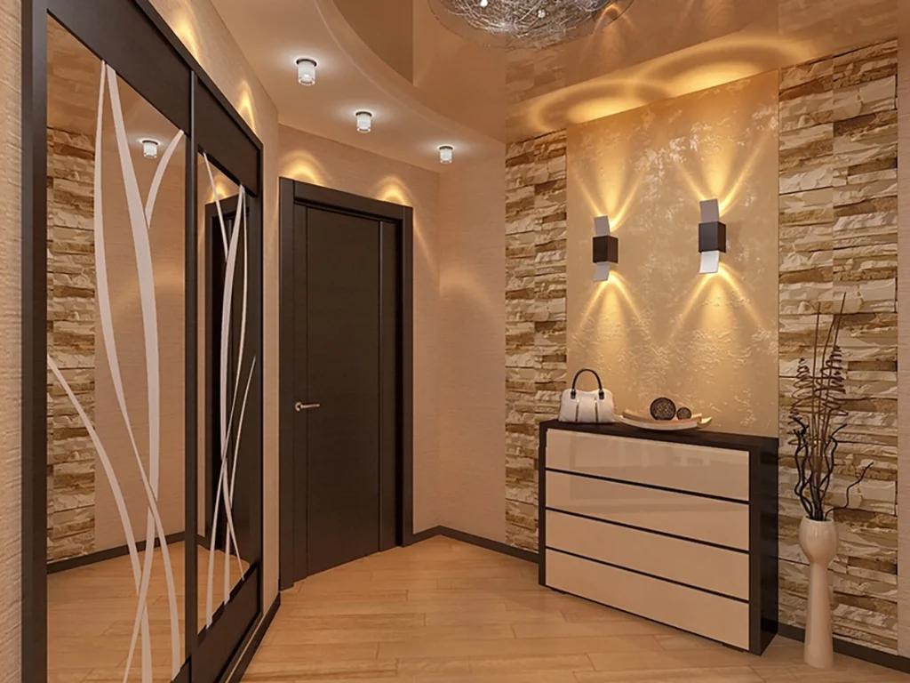 A modern home hallway featuring warm ambient lighting from sleek wall sconces and recessed ceiling lights. An accent wall with stone brick detailing frames a glossy beige chest of drawers, while a large sliding wardrobe with frosted glass leaf patterns stands on the left.