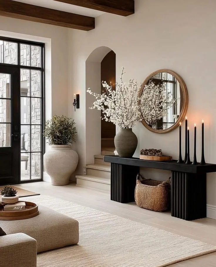 A bright, open-concept modern organic entryway featuring a chunky black console table with fluted legs, styled with tall white cherry blossom branches in a textured vase and elegant black taper candles.