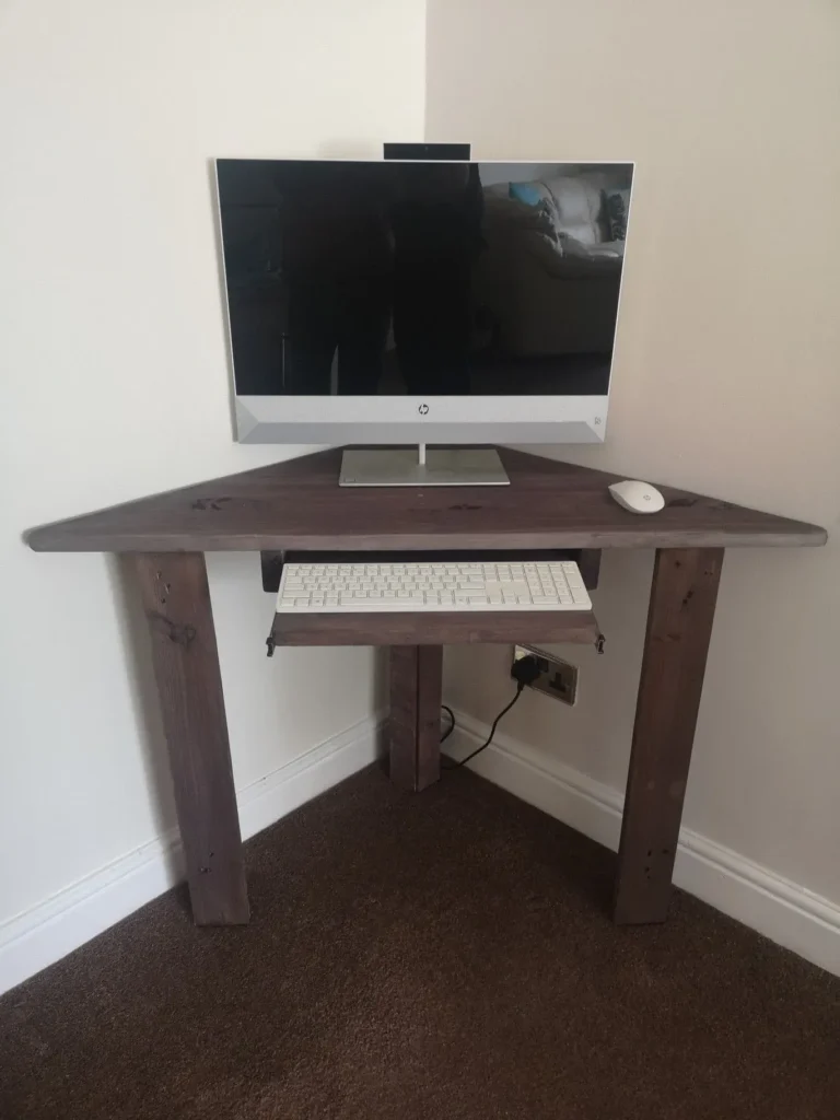A dark, rustic wooden triangular corner desk tucked into a room corner with beige walls and brown carpet. An HP all-in-one computer monitor sits on the desktop next to a white wireless mouse. A slide-out keyboard tray underneath holds a white computer keyboard, and a power cord plugs into a wall outlet under the desk.