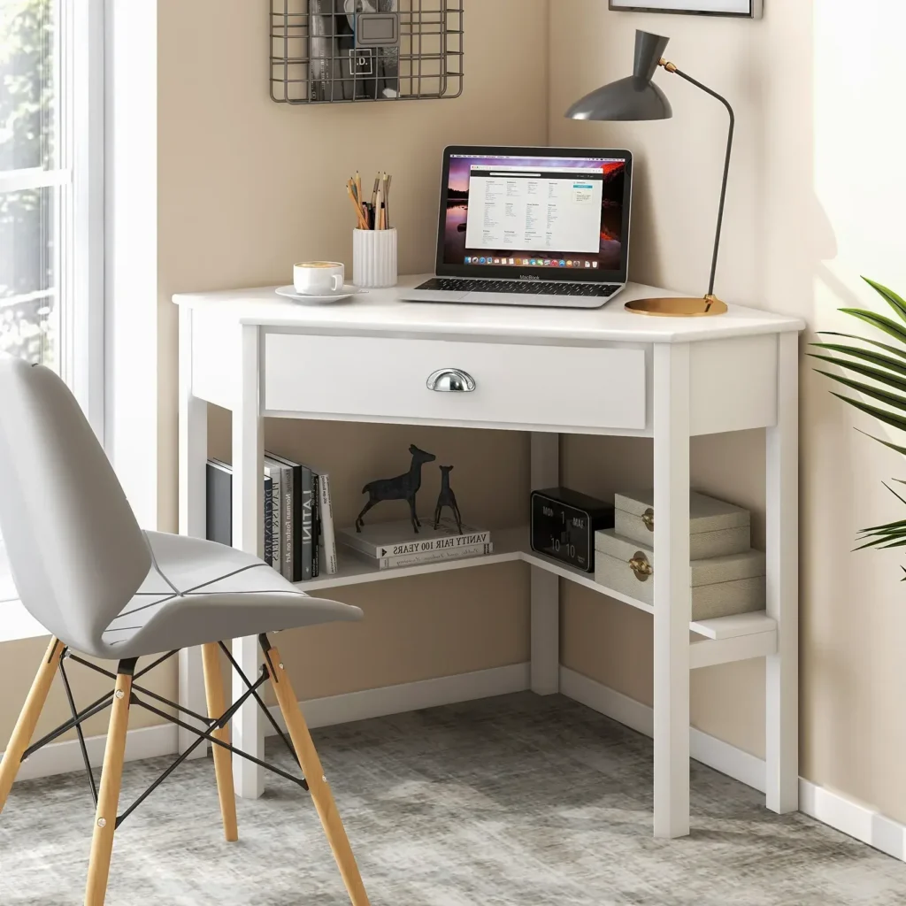 Space-saving white corner writing desk tucked into a room corner, featuring a sleek silver drawer handle, open under-desk shelving, and a laptop setup.