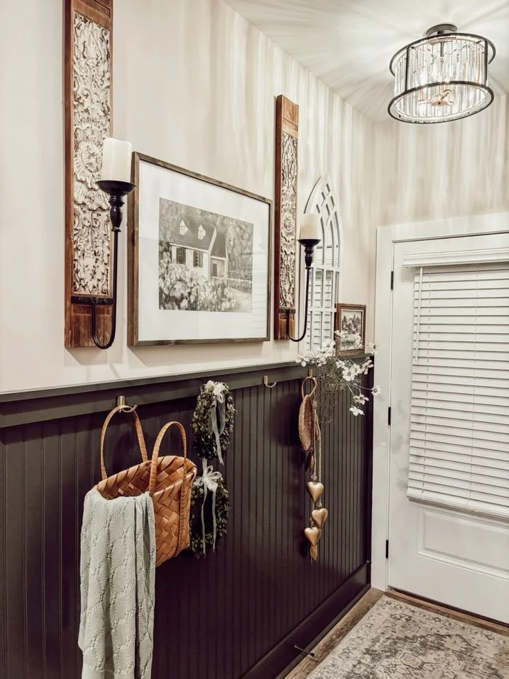 A small farmhouse-style entryway featuring dark gray beadboard wainscoting with a peg rail. Framed black-and-white artwork of a house hangs on the wall above, flanked by two rustic wooden wall plaques with black metal candle sconces. A woven market basket with a blanket, small green wreaths, and dried flowers hang from the metal wall hooks. A classic white front door with window blinds sits adjacent to the decorated wall under a modern crystal drum chandelier.
