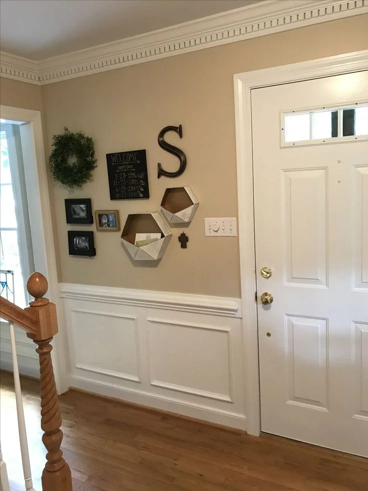A small entryway with beige walls, a classic white front door, and white wainscoting paneling along the lower wall. A personalized gallery wall features a large metal letter "S," two white hexagonal geometric wall pockets holding mail, a small chalkboard with family dates, a green wreath, and framed family photos. The space has light hardwood floors and detailed crown molding, with a wooden staircase banister visible in the foreground.