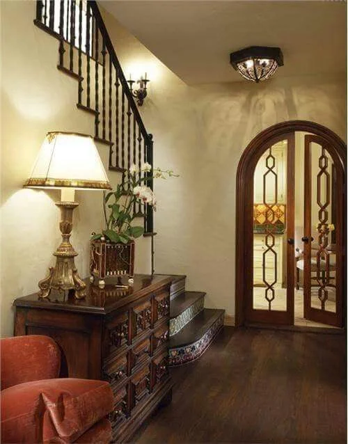 A traditional small foyer featuring a dark wood chest of drawers placed next to a staircase with ornate black metal railings and decorative tiled risers. An elegant, classic table lamp with a large cream shade and a potted white orchid sit on top of the chest. The entryway opens into the next room through arched, dark wood double glass doors with a geometric fretwork pattern over dark hardwood flooring.