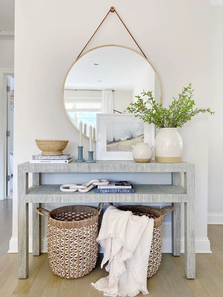 A bright, modern small foyer featuring a light gray textured linen console table with a built-in lower shelf. Hanging above the table is a large round mirror suspended by a tan leather strap. The tabletop is styled with a large white ceramic vase filled with bright green leafy branches, a smaller two-tone vase, a framed landscape art piece, two blue taper candle holders, and a woven decorative bowl stacked on books. On the bottom floor space, two woven storage baskets are placed side-by-side, with a light pink fringed throw blanket draped over one.