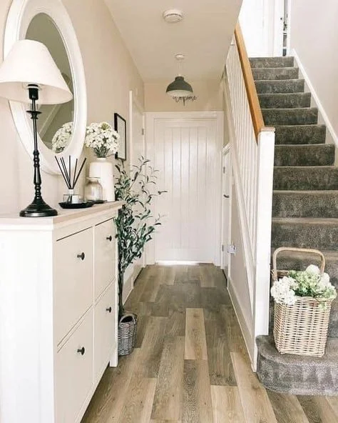 narrow hallway shoe storage ideas A narrow home entryway featuring a slim white shoe storage cabinet with a table lamp and decor on top, a large white round mirror, wood-look flooring, and a carpeted staircase with a decorative wicker basket on the bottom step.