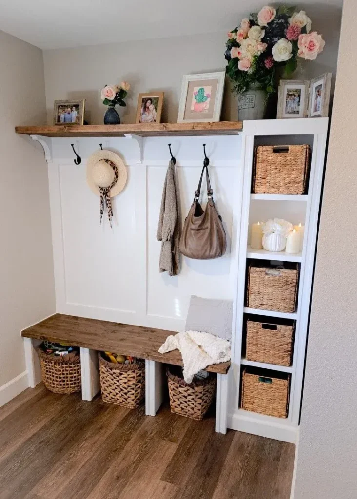 built in hallway mudroom bench ideas A custom white built-in mudroom unit featuring a dark wood bench and top shelf, black coat hooks holding a purse and hat, open cubbies with woven storage baskets, and a tall side shelving tower.