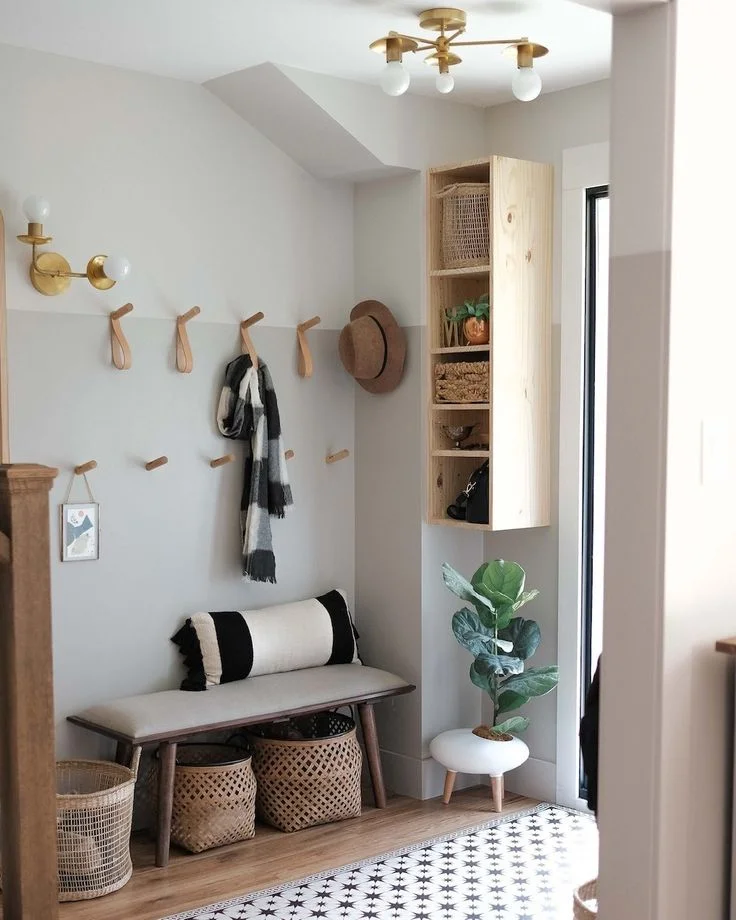 Tiny Entryway Storage Makeover - Woven Baskets, Wood Hooks, and Shelf Unit in small space. A small, stylish entryway optimized for storage, featuring two-tone grey walls adorned with light-wood hooks, a modern upholstered wooden bench with a black-and-white bolster pillow, and several woven storage baskets. A wall-mounted multi-tiered wooden shelf unit and a faux fiddle-leaf fig tree sit to the right, all illuminated by warm brass light fixtures.
