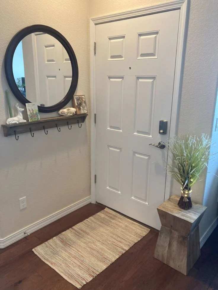 Tiny Entryway Makeover with Rustic Shelf and Wood Pedestal. A tiny entryway featuring dark wood floors, a patterned rug, a white paneled front door with a smart lock, a round black-framed mirror above a rustic wooden floating shelf with hooks, and a wooden pedestal holding a bundle of lighted dried grass.