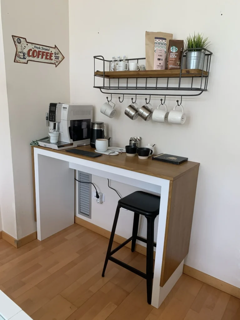 Organized DIY coffee corner showcasing a modern espresso machine on a white and wood counter table, a single black stool, and an overhead rack displaying ceramic mugs and coffee beans.
