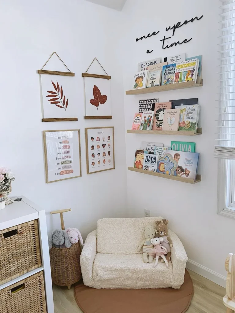 Organized small space toddler reading corner with a plush white accent chair, three wooden floating book ledges on a white wall, and neutral framed posters featuring leaves and positive affirmations.