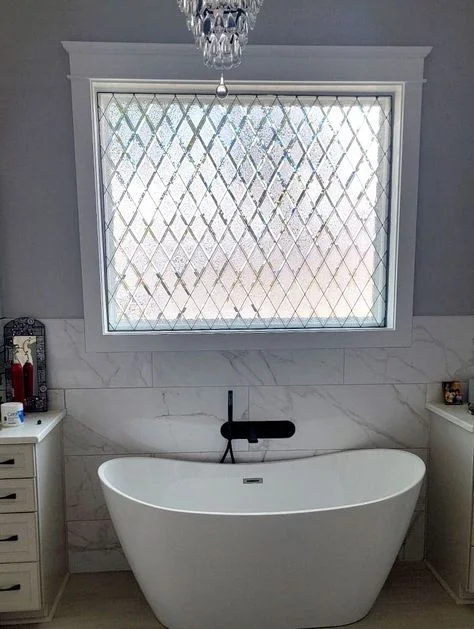 "An elegant bathroom featuring a freestanding white soaking tub with a modern black wall-mounted faucet, set against a white marble-tiled wall. Above the tub, a white-framed window with a textured, diamond-patterned leaded glass design provides privacy under a hanging crystal chandelier."