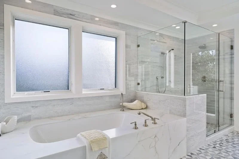 "A bright, upscale master bathroom featuring a built-in bathtub surrounded by white marble. Two large white-framed windows with frosted privacy glass sit directly above the tub, next to a modern walk-in shower with a frameless glass enclosure."