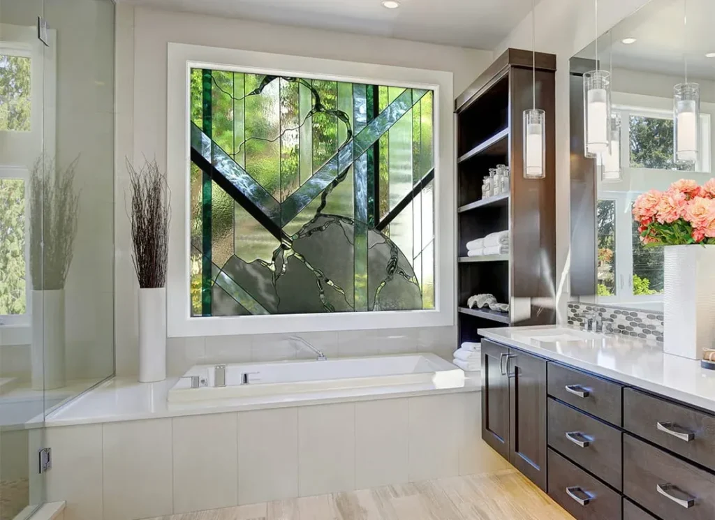 A wide-angle view of a modern master bathroom showing a large white soaking tub directly underneath a white-framed window. The window features an intricate, abstract stained glass design in shades of green, black, and grey, ensuring privacy while acting as a focal point. To the right is a dark wood double-sink vanity and to the left is a frameless glass shower enclosure.