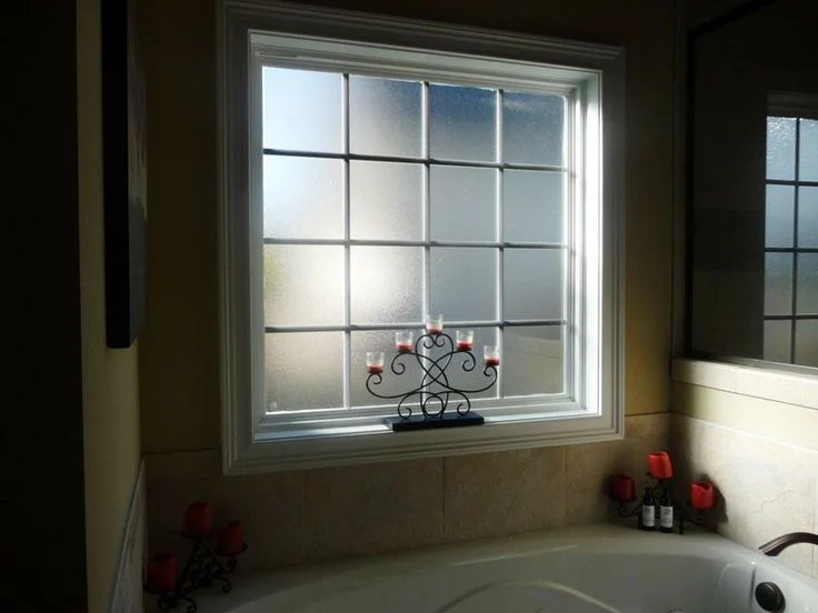 A square, gridded bathroom window featuring textured frosted glass for privacy, positioned directly above a corner bathtub decorated with elegant black metal candelabras.