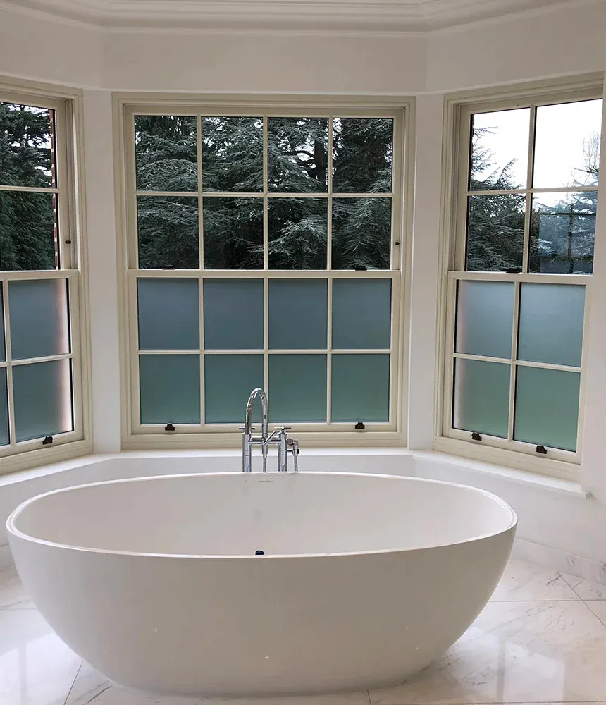 A large white freestanding soaking tub is centered in front of a three-part bay window. Each casement window features frosted glass on the lower panes, providing direct privacy for the bather, while the clear upper panes offer a view of a mature pine tree forest outside.