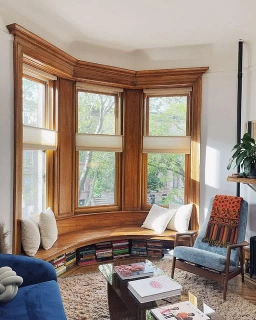 A cozy living room reading nook featuring a large bay window with rich natural wood framing and a matching built-in curved wooden bench. The space underneath the bench is filled with rows of stacked books, creating a clever bookshelf display. The scene is completed by a shaggy beige area rug, a glass coffee table, a mid-century blue armchair draped with an orange patterned throw blanket, and neutral accent pillows.