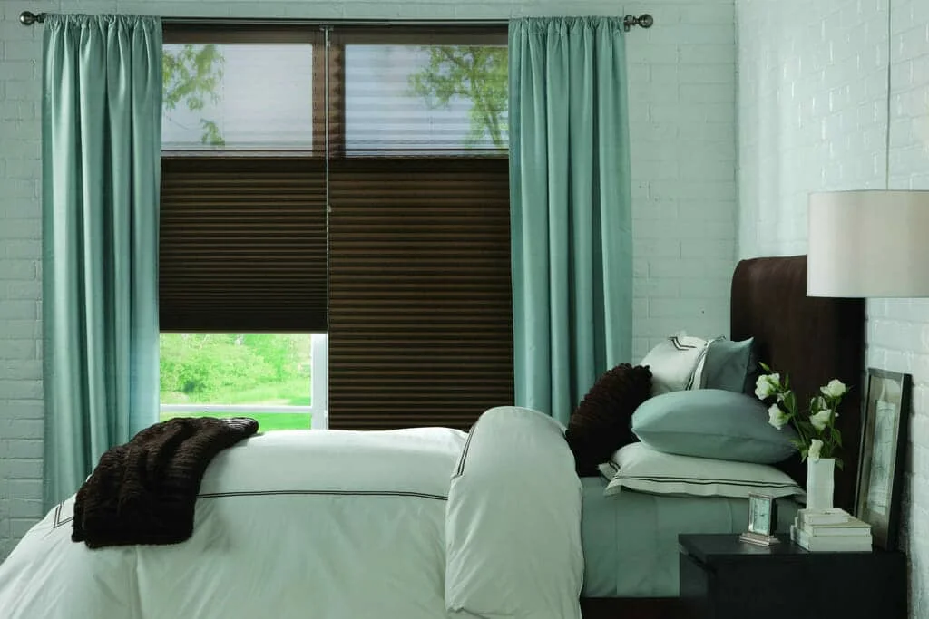 Modern bedroom window featuring dark brown top-down bottom-up cellular shades framed by floor-length mint green drapes, with a dark brown upholstered headboard and light green bedding.