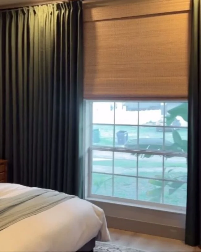 Bedroom window featuring a natural warm-toned woven Roman shade paired with floor-length forest green curtains beside a bed with white and sage green bedding.