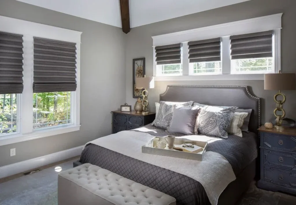 A photograph of a well-designed bedroom showcasing dark grey Roman shades on the windows, a grey leather headboard, patterned pillows, a breakfast tray on the bed, and a tufted ottoman at the foot of the bed.