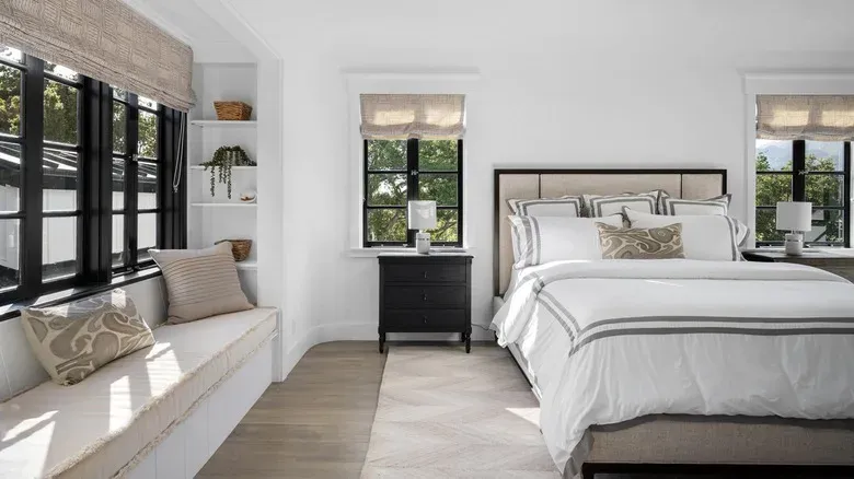 Bright white bedroom featuring neutral fabric Roman shades on black framed windows, a built-in window seat, and a upholstered bed with white linens.