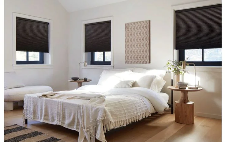 Dark grey honeycomb window shades partially lowered in a sunlit white bedroom with neutral bedding and natural wood accents.