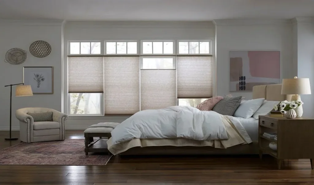 Spacious neutral bedroom featuring a bank of large windows fitted with taupe top-down bottom-up cellular shades, an upholstered bed with white bedding, and a beige armchair nook.
