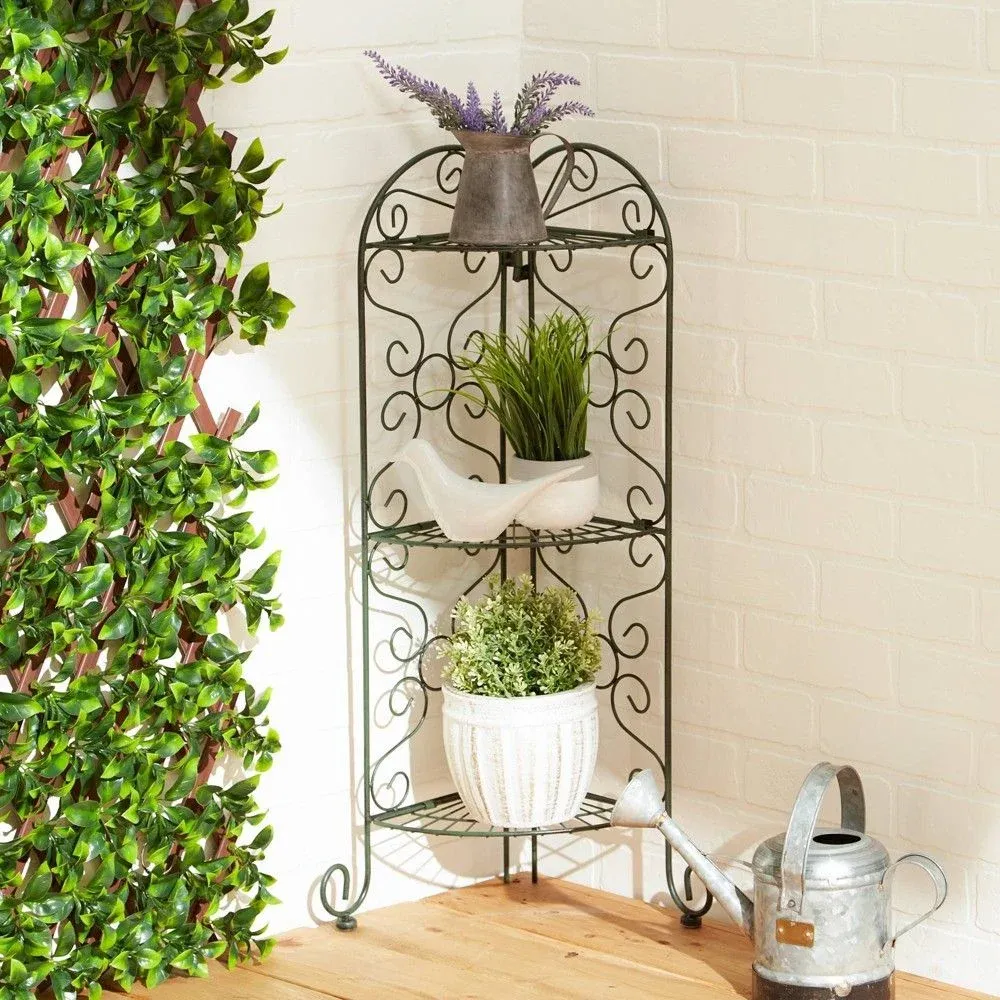 Wrought iron style three-tier corner plant display stand featuring potted greenery, a decorative bird figurine, and a metal watering can against a white brick background.